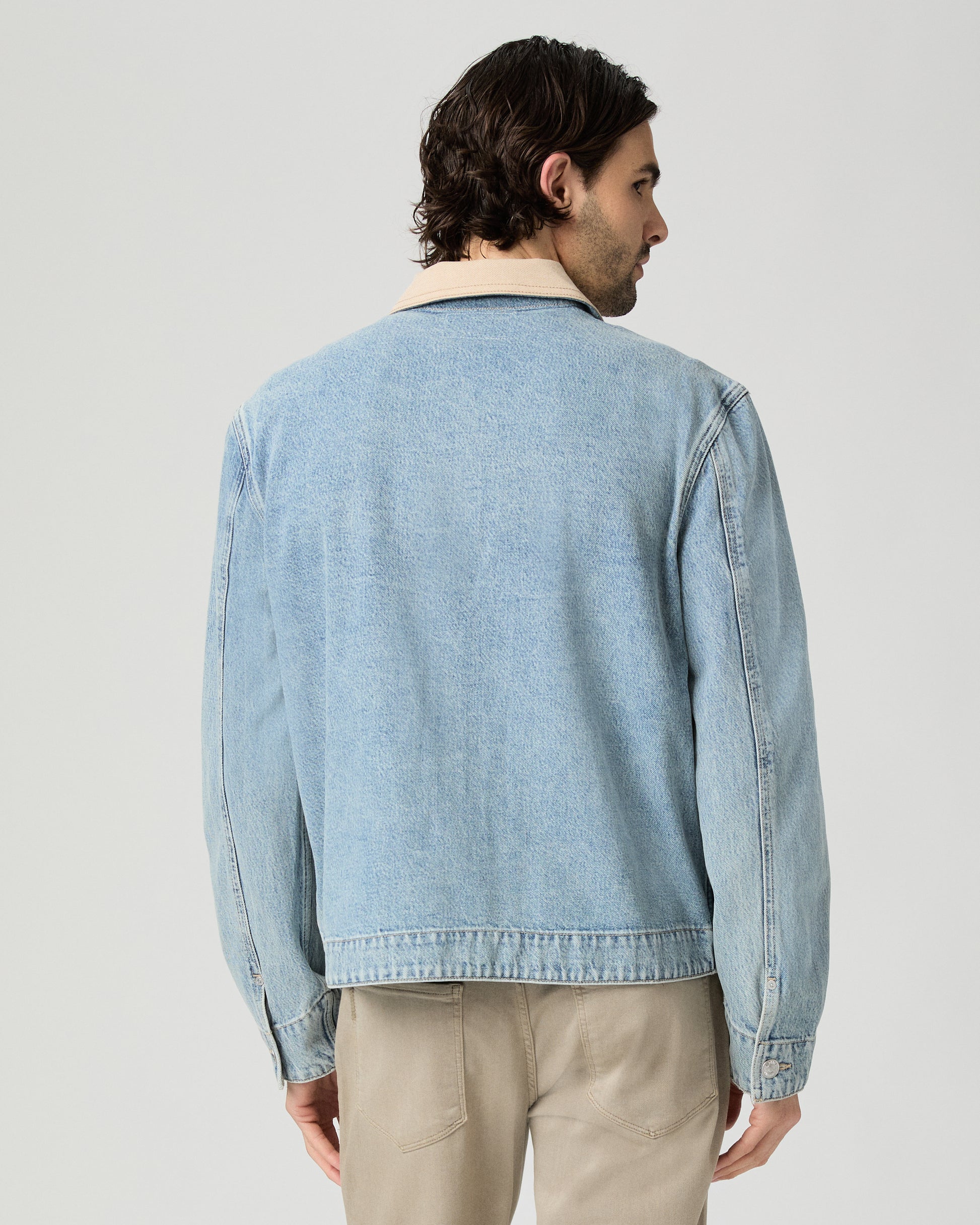 Man wearing a light blue denim jacket over a white shirt and beige pants on a gray background