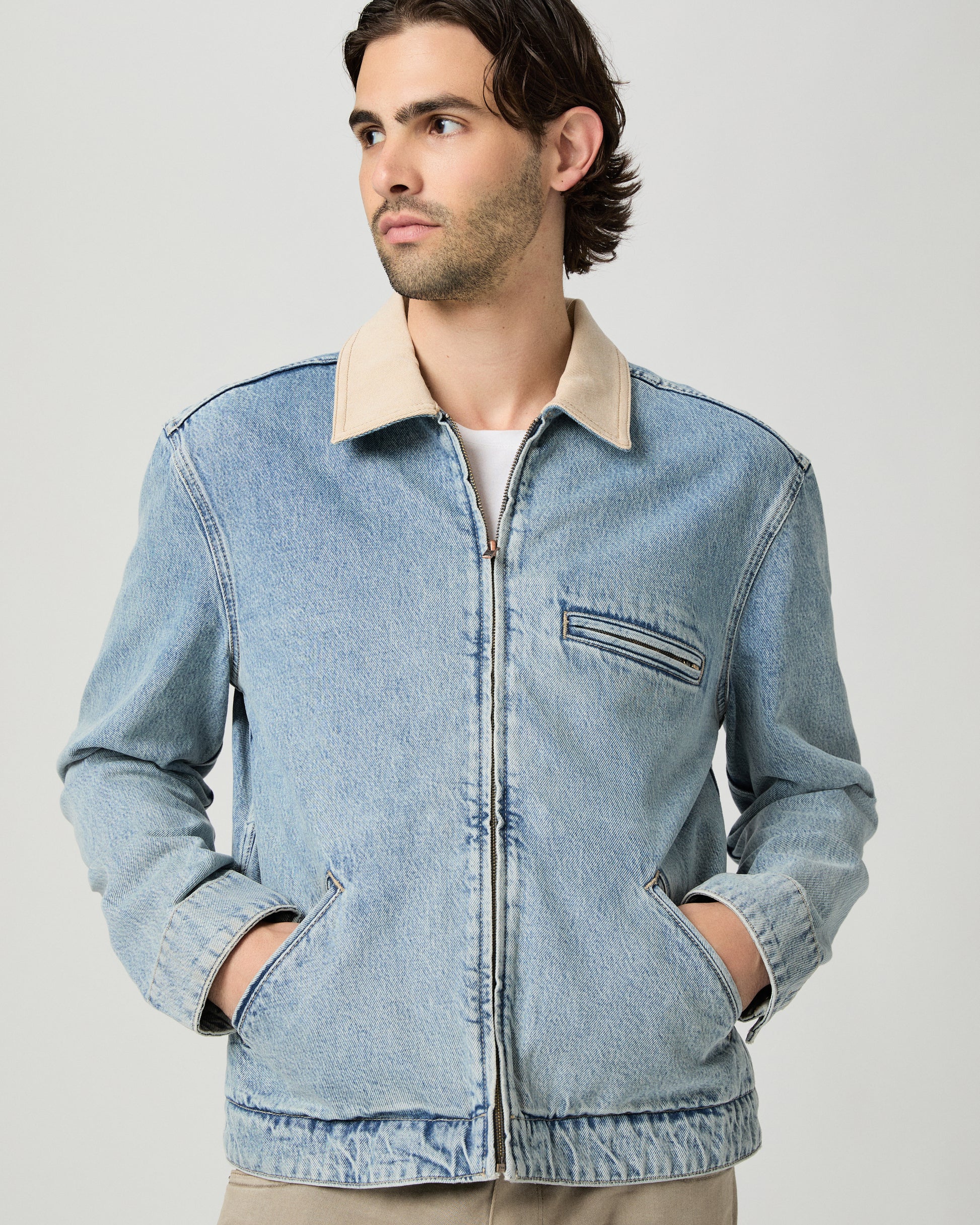 Man wearing a light blue denim jacket over a white shirt and beige pants on a gray background
