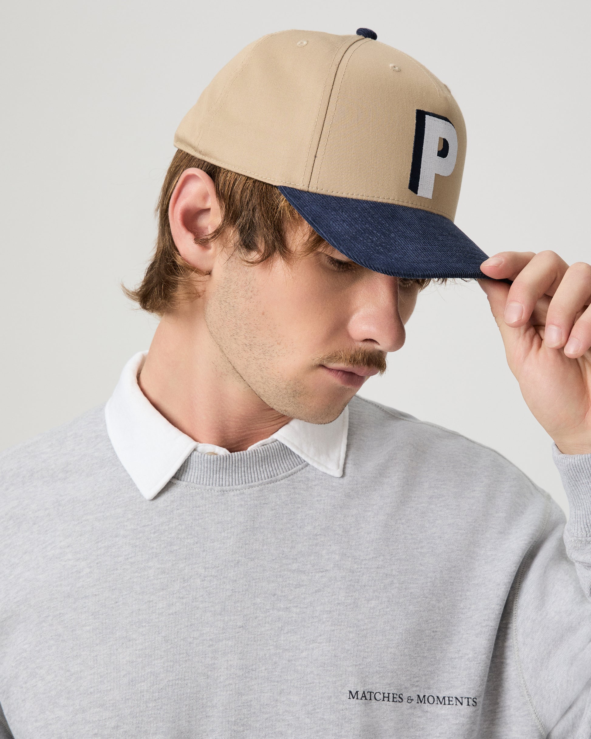 Man wearing a beige and navy cap with a logo, looking down, on a plain background.