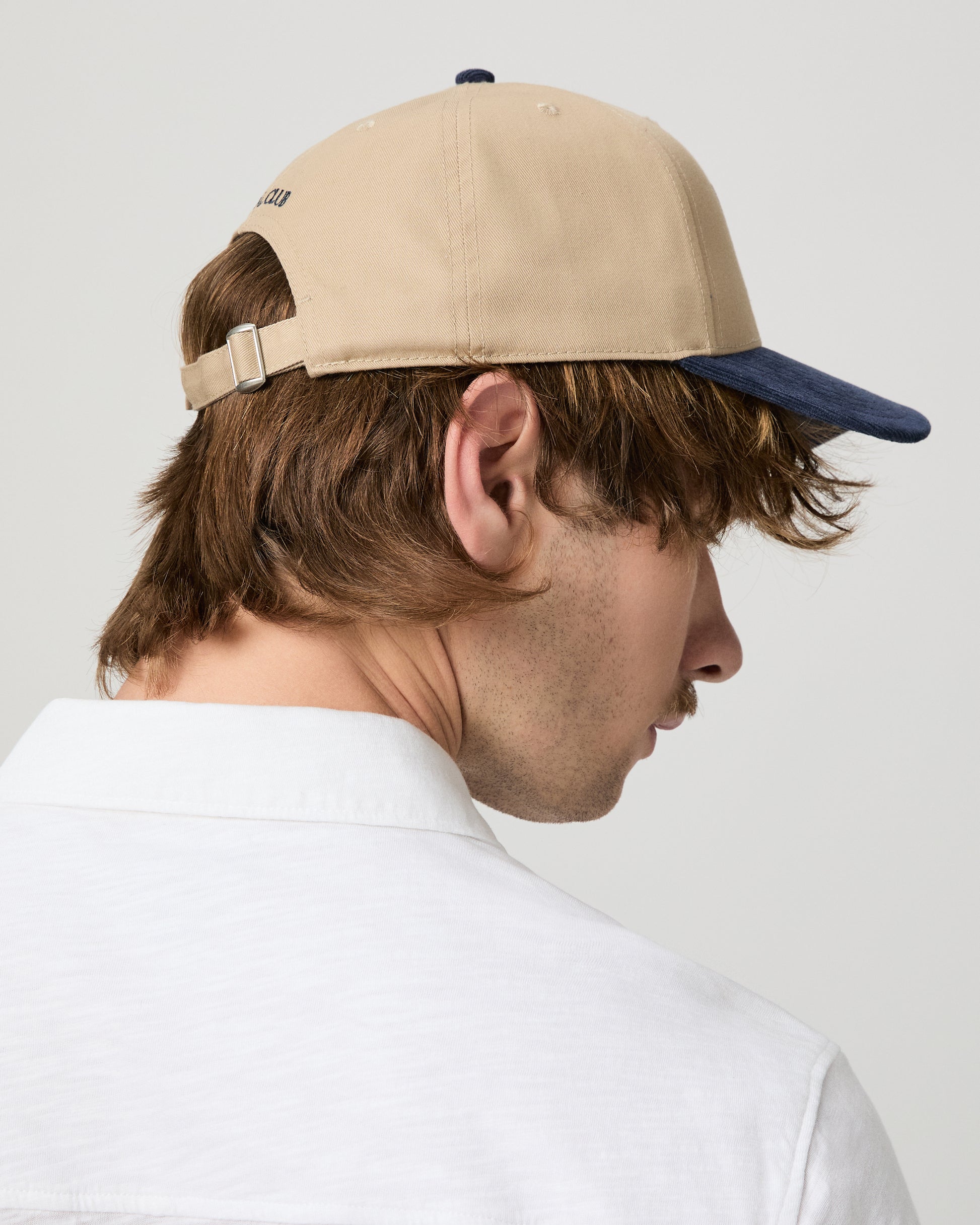 Man wearing a beige and navy cap with a logo, looking down, on a plain background.
