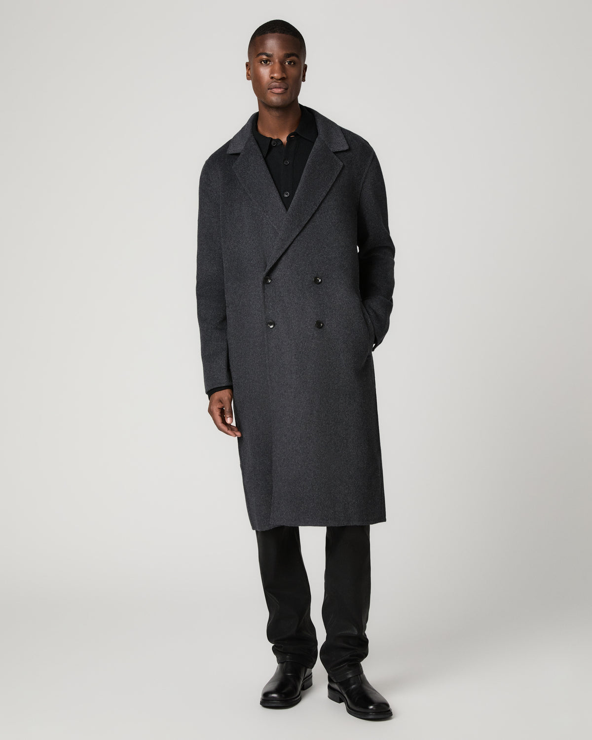 Full-length front view of a man with one hand in his pocket, wearing the dark charcoal double-breasted Anselmo Coat with notched lapels over a black polo shirt, black trousers, and black boots.