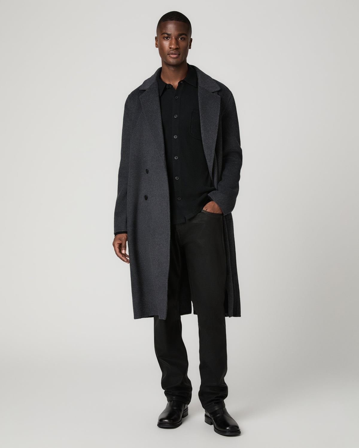 Front view of a man wearing the dark charcoal double-breasted Anselmo Coat open and unbuttoned over an all-black shirt and trousers, with one hand tucked in his pocket.