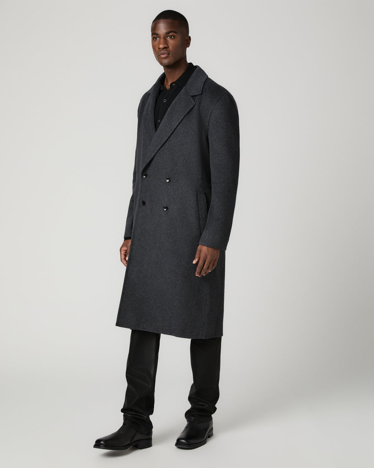 Three-quarter angle view of a man wearing the dark charcoal double-breasted Anselmo Coat buttoned closed over a black polo shirt, paired with black trousers and boots.
