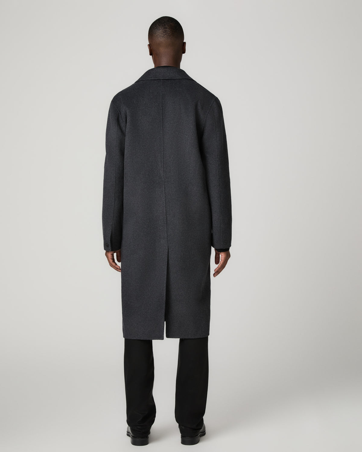 Back view of a man wearing the full-length dark charcoal Anselmo Coat, showing the back vent and clean tailored silhouette over black trousers.