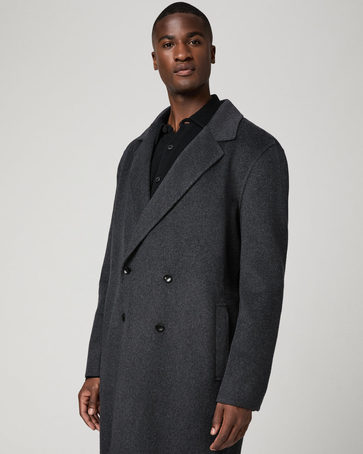 Close-up view of a man wearing the dark charcoal double-breasted Anselmo Coat, highlighting the notched lapel and button detail over a black polo shirt.