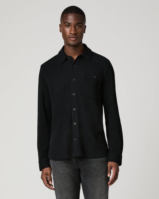 Cropped front view of a man wearing a black thermal waffle-knit button-down shirt with a chest pocket, tucked into gray denim jeans.