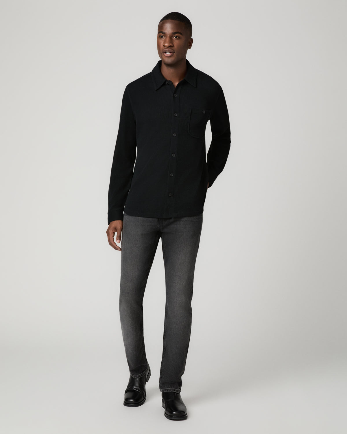 Full-length front view of a man wearing a black waffle-knit shirt buttoned to the collar, tucked into gray skinny jeans with black leather shoes.