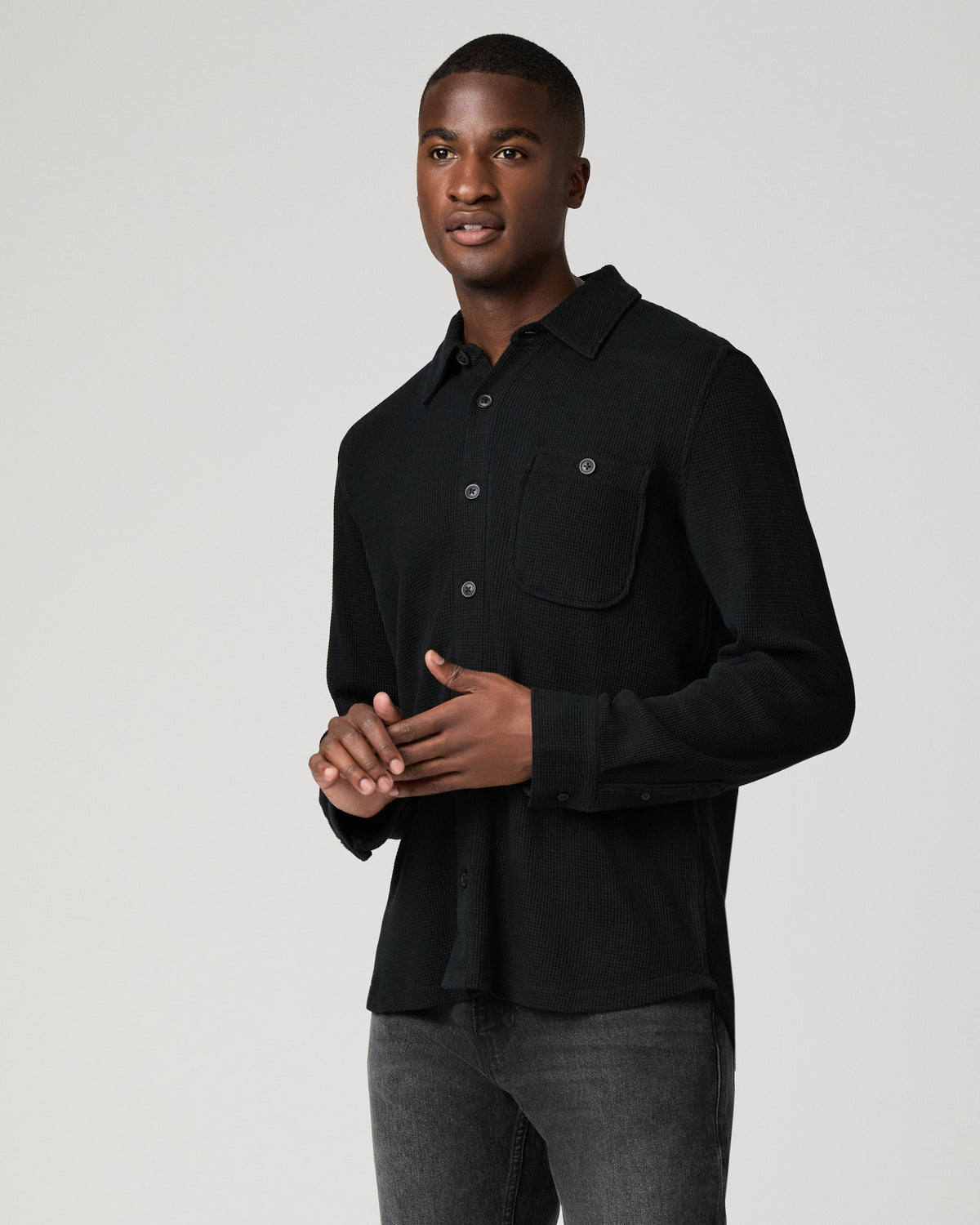 Three-quarter view of a man in a black waffle-knit shirt with hands clasped near his chest, highlighting the chest pocket and tonal button detail.