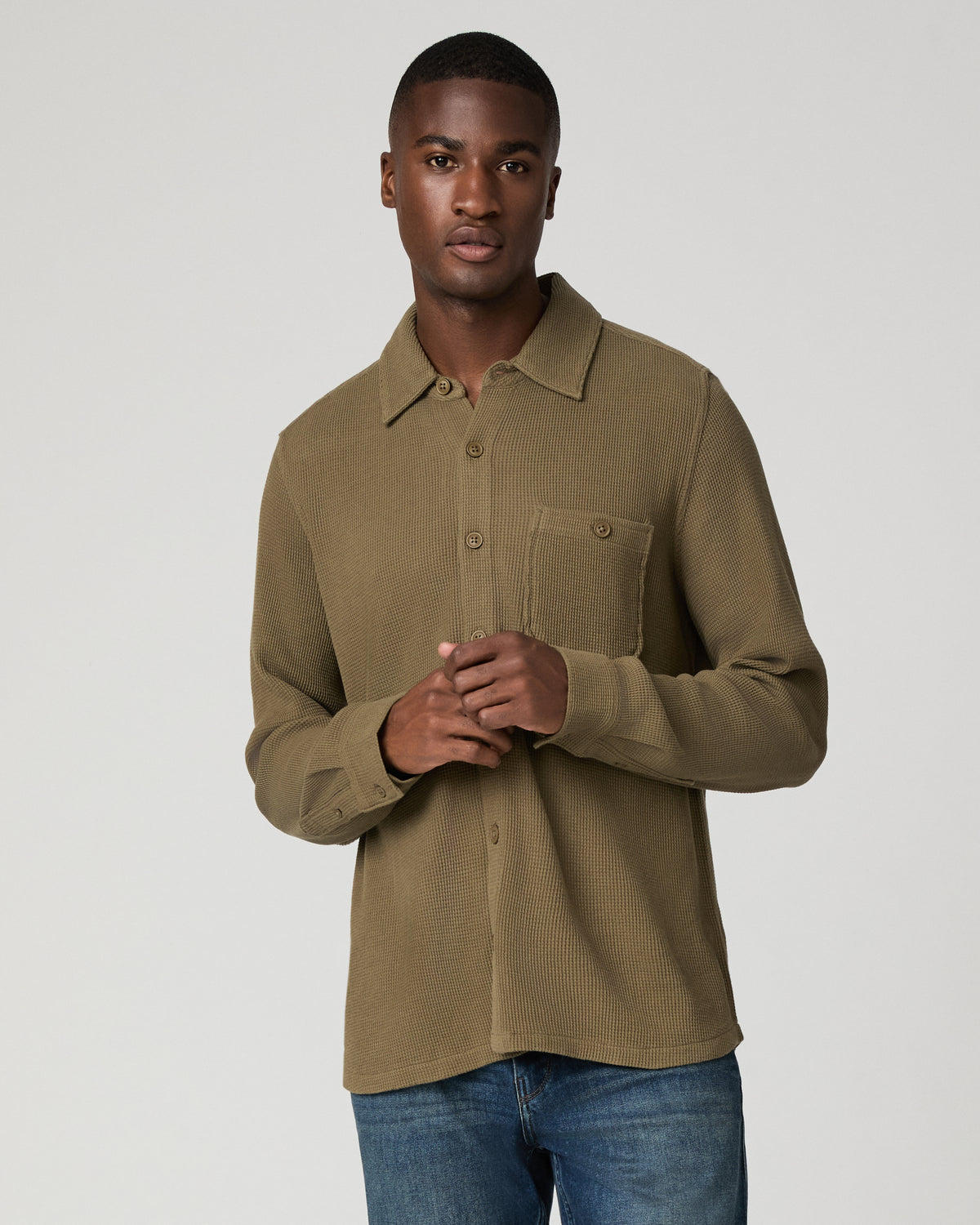 Cropped front view of a man with hands clasped wearing a deep olive green waffle-knit button-down shirt with a chest pocket, tucked into blue denim jeans.