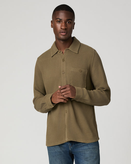 Cropped front view of a man with hands clasped wearing a deep olive green waffle-knit button-down shirt with a chest pocket, tucked into blue denim jeans.