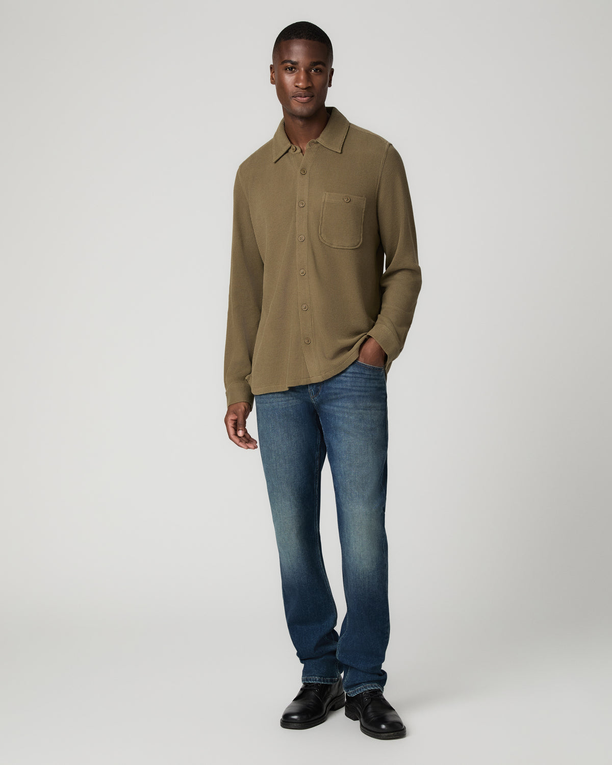 Full-length front view of a man wearing a deep olive green waffle-knit shirt buttoned to the collar, tucked into blue denim jeans with black leather shoes, one hand in his pocket.