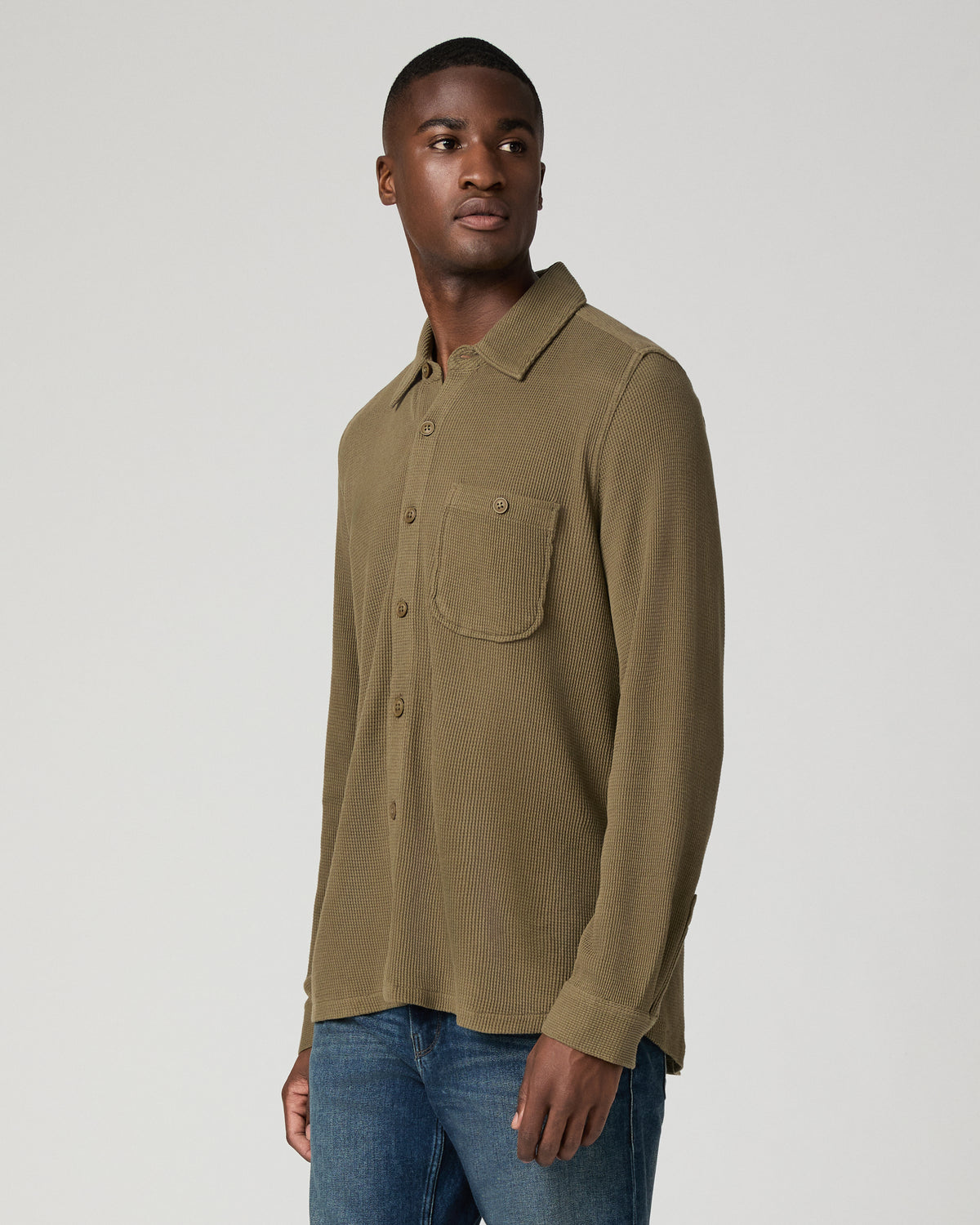 Three-quarter side view of a man in a deep olive green waffle-knit shirt, highlighting the chest pocket and tonal button detail against blue jeans.