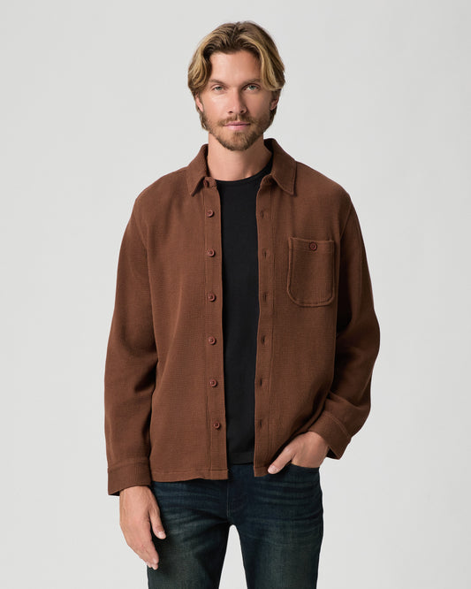 Model standing with one hand in his pocket, wearing the Hollander Shirt in Sepia Clay, a brown button-front shirt worn open over a black tee, with dark jeans