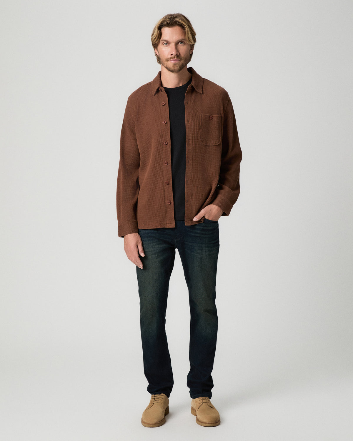 Full-length view of model with one hand in his pocket, wearing the Hollander Shirt in Sepia Clay open over a black tee with dark-wash jeans and tan suede boots