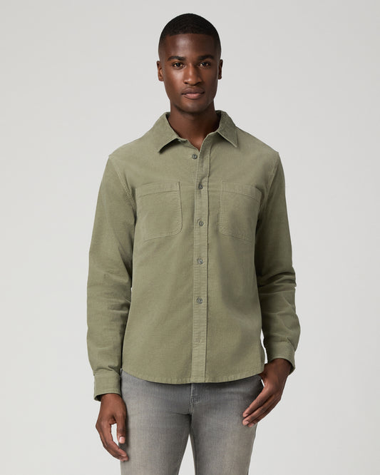 Cropped front view of a man wearing a sage green corduroy button-down shirt with a chest pocket, tucked into gray denim jeans.