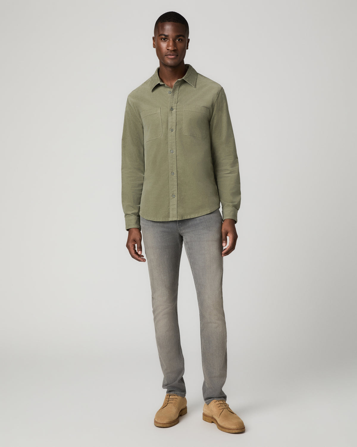 Full-length front view of a man wearing a buttoned sage green corduroy shirt tucked into gray skinny jeans with tan suede chukka boots.