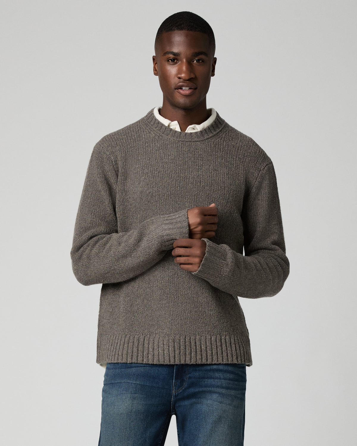 Front view of a man wearing a dark heather grey ribbed crewneck sweater layered over a white collared shirt, paired with dark wash jeans, hands clasped in front of him.