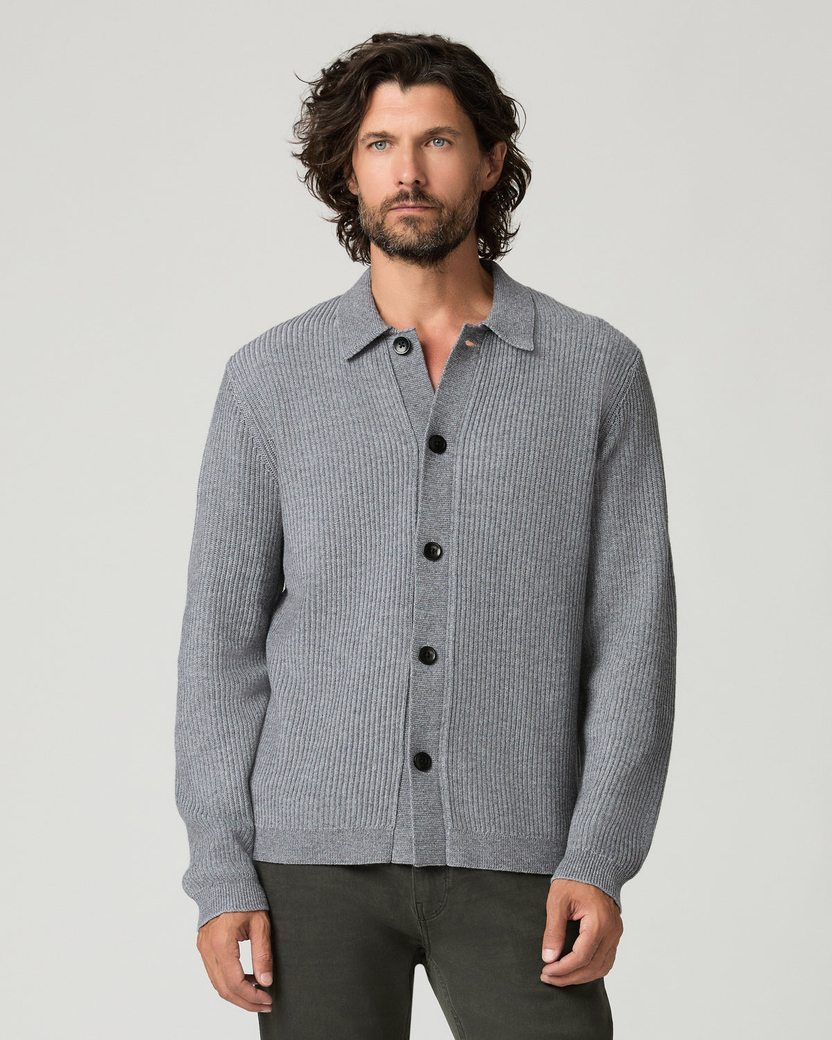 Front view of a man standing with arms crossed, wearing the heather gray ribbed-knit Spokane Cardigan buttoned to the collar over dark olive trousers and black leather shoes.
