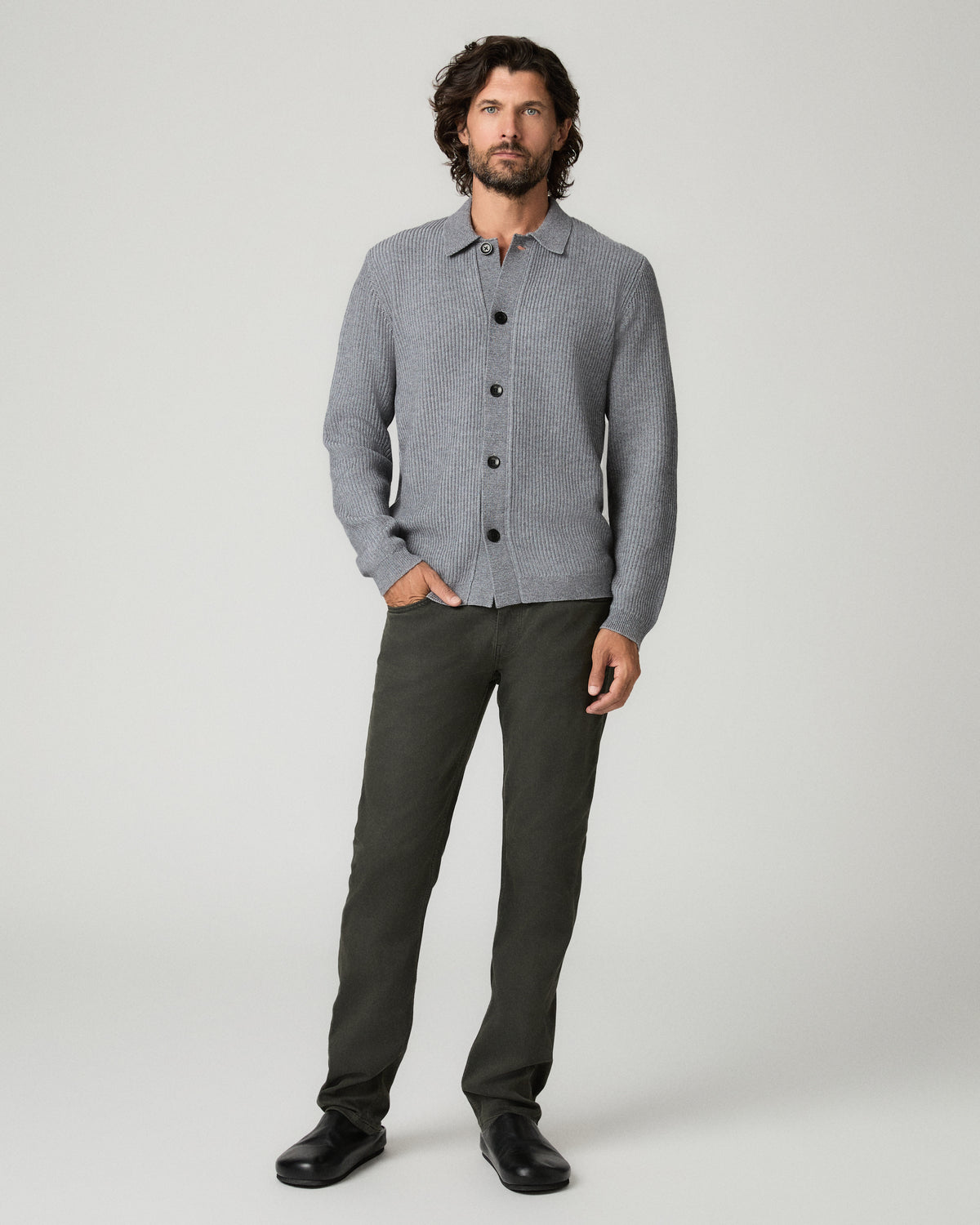 Full-length front view of a man with one hand in his pocket, wearing the heather gray ribbed-knit Spokane Cardigan fully buttoned over a white tank top, paired with dark olive trousers and black shoes.