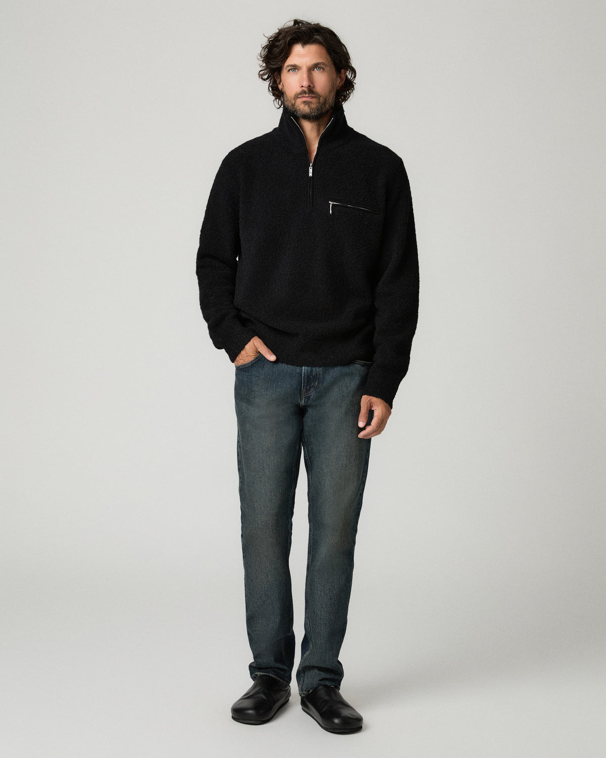 Full-length front view of a man standing with one hand in his pocket, wearing the black bouclé-knit Donover Quarter Zip Sweater over dark straight-leg jeans and black shoes.