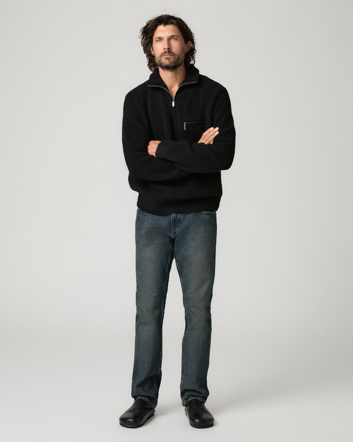 Front view of a man standing with his arms crossed, wearing the black bouclé-knit Donover Quarter Zip Sweater with the collar zipped high, paired with dark jeans and black shoes.