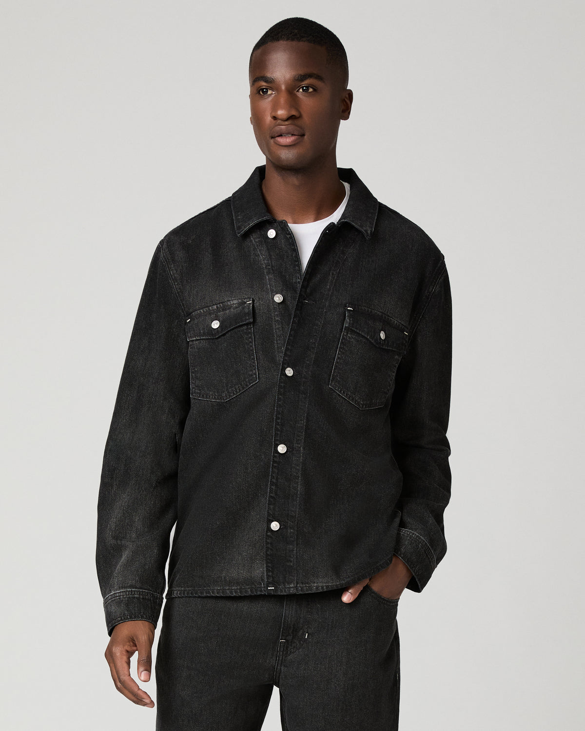 Cropped front view of a man wearing a charcoal wash denim shirt with snap-button closures and flap chest pockets, one hand tucked into his matching dark denim trousers.