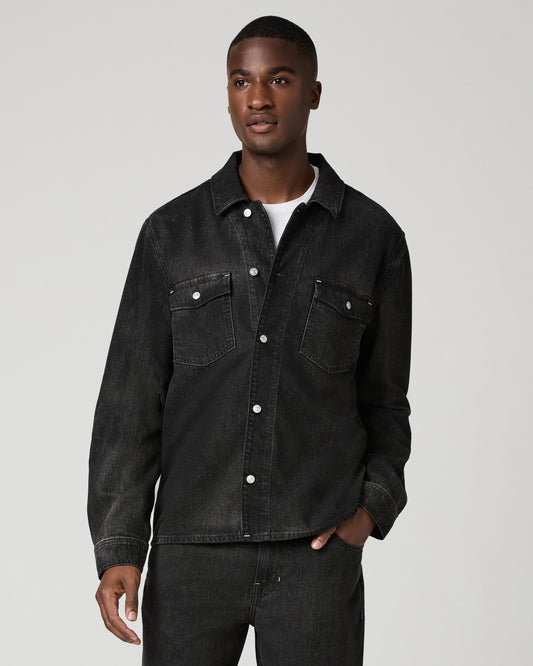 Cropped front view of a man wearing a charcoal wash denim shirt with snap-button closures and flap chest pockets, one hand tucked into his matching dark denim trousers.