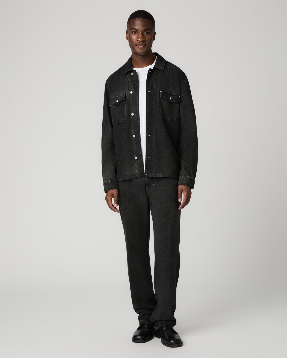 Full-length front view of a man wearing an open-collar charcoal denim shirt over a white tee, tucked into slim dark jeans with black leather shoes.