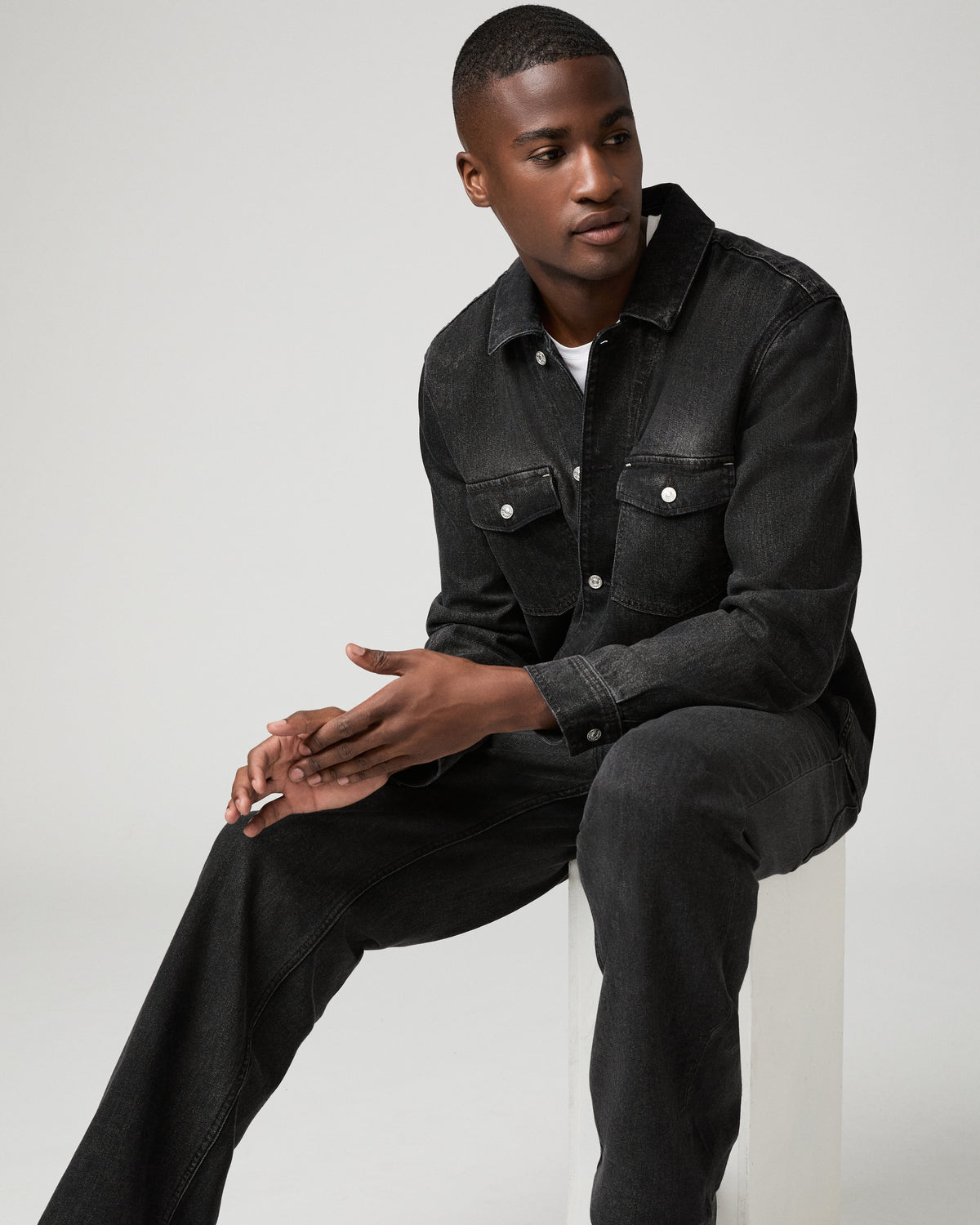 Seated front view of a man wearing a buttoned charcoal denim shirt with a white tee underneath, hands clasped in his lap.