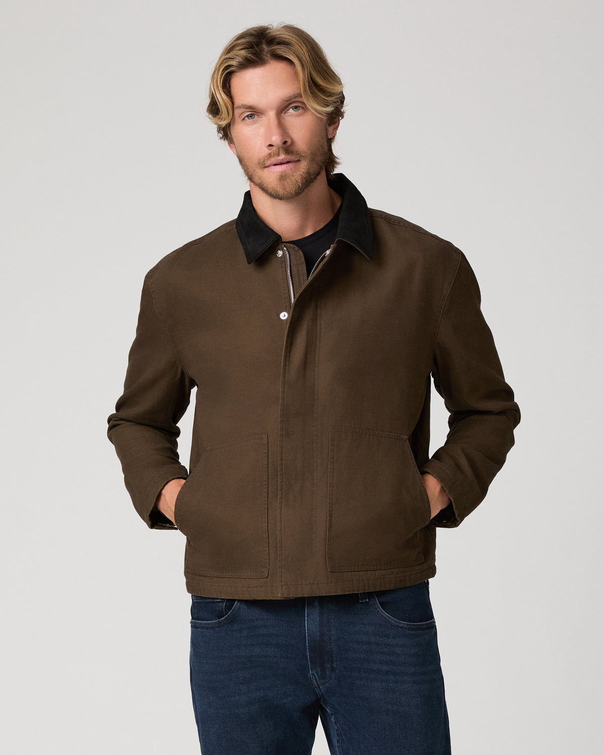 A man wearing a brown waxed cotton utility jacket with a black collar, paired with dark blue jeans, photographed against a neutral light gray studio background.