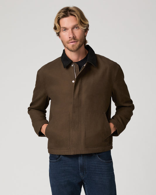 A man wearing a brown waxed cotton utility jacket with a black collar, paired with dark blue jeans, photographed against a neutral light gray studio background.