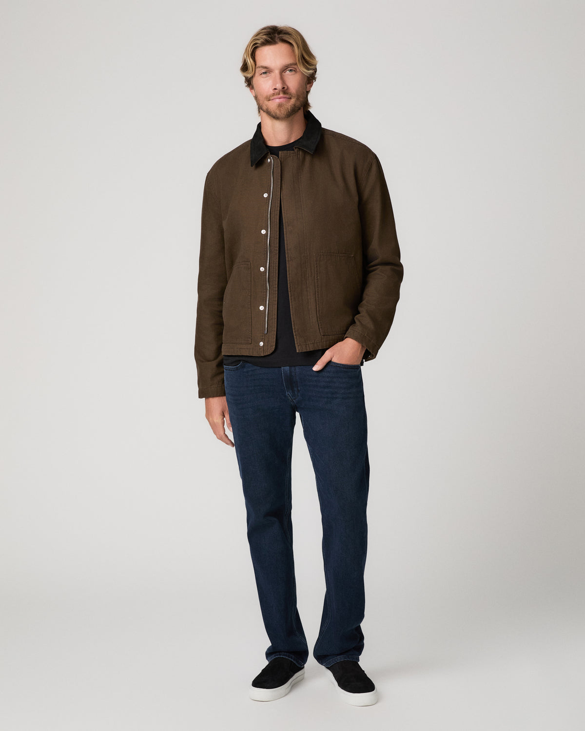 A man wearing a brown waxed cotton utility jacket with a black collar, paired with dark blue jeans and black shoes photographed against a neutral light gray studio background.