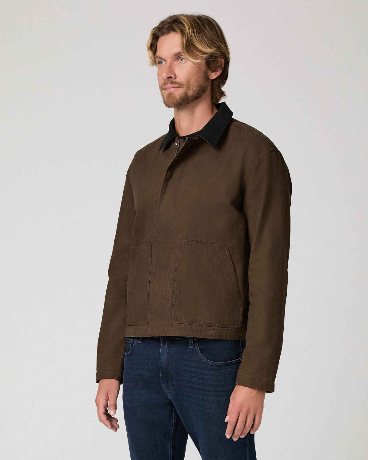 A man wearing a brown waxed cotton utility jacket with a black collar, paired with dark blue jeans, photographed against a neutral light gray studio background.