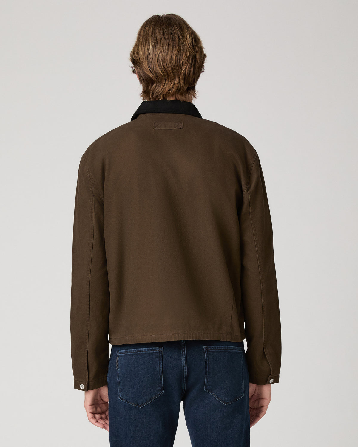 A man wearing a brown waxed cotton utility jacket with a black collar, paired with dark blue jeans, photographed against a neutral light gray studio background.