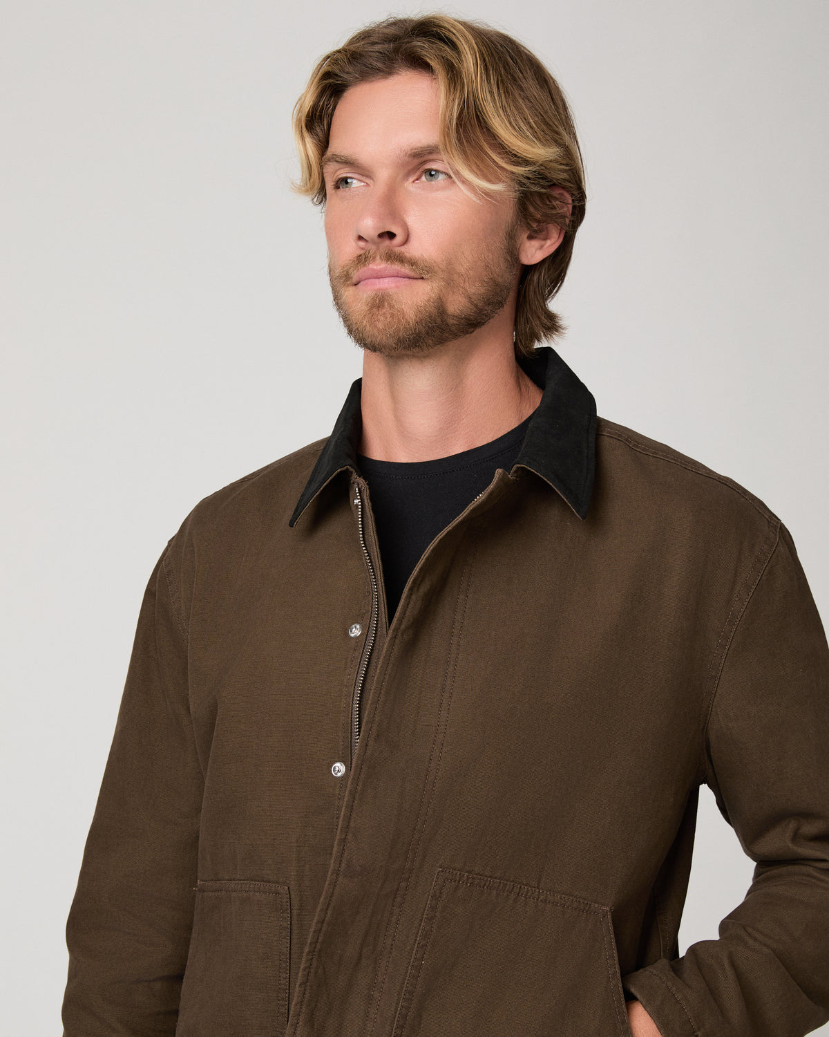 A man wearing a brown waxed cotton utility jacket photographed against a neutral light gray studio background.