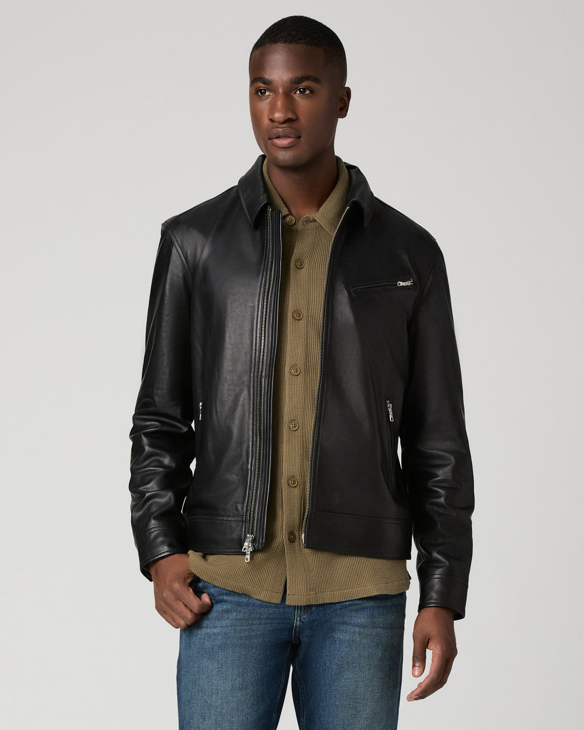 Front view of a man wearing the black leather Rocoso Moto Jacket unzipped over an olive button-up shirt, paired with blue jeans, showing the point collar and silver-tone zip hardware.