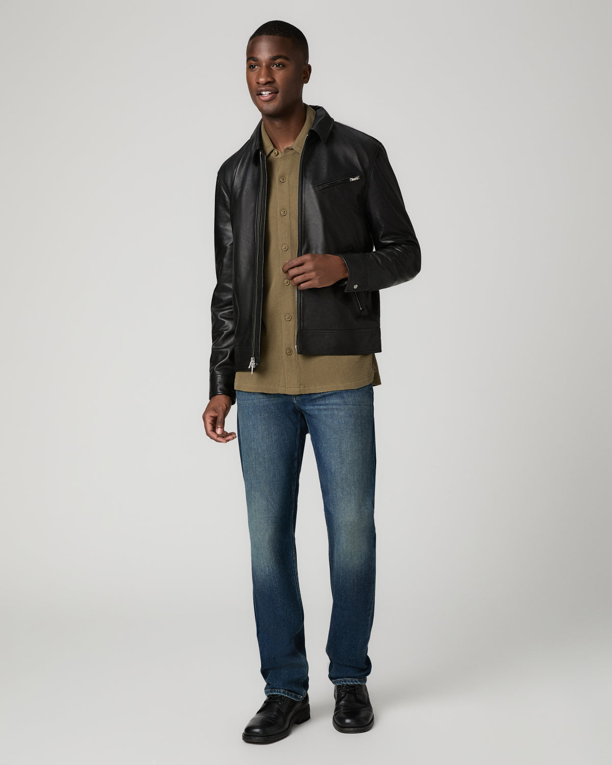 Full-length front view of a man walking, one hand touching the lapel of the black leather Rocoso Moto Jacket, worn over an olive shirt with blue jeans and black shoes.