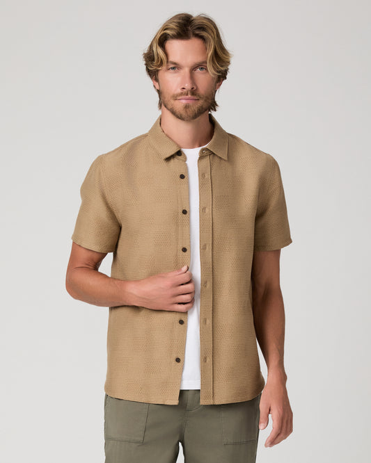 A man wearing a tan short-sleeve button-up shirt over a white t-shirt and olive green pants, standing against a light gray studio background.