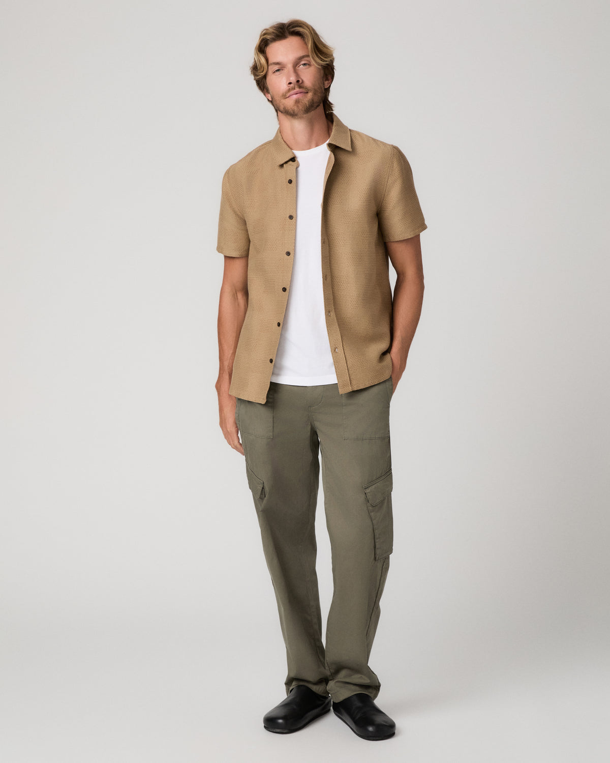 A man wearing a tan short-sleeve button-up shirt over a white t-shirt and olive green pants and black shoes, standing against a light gray studio background.