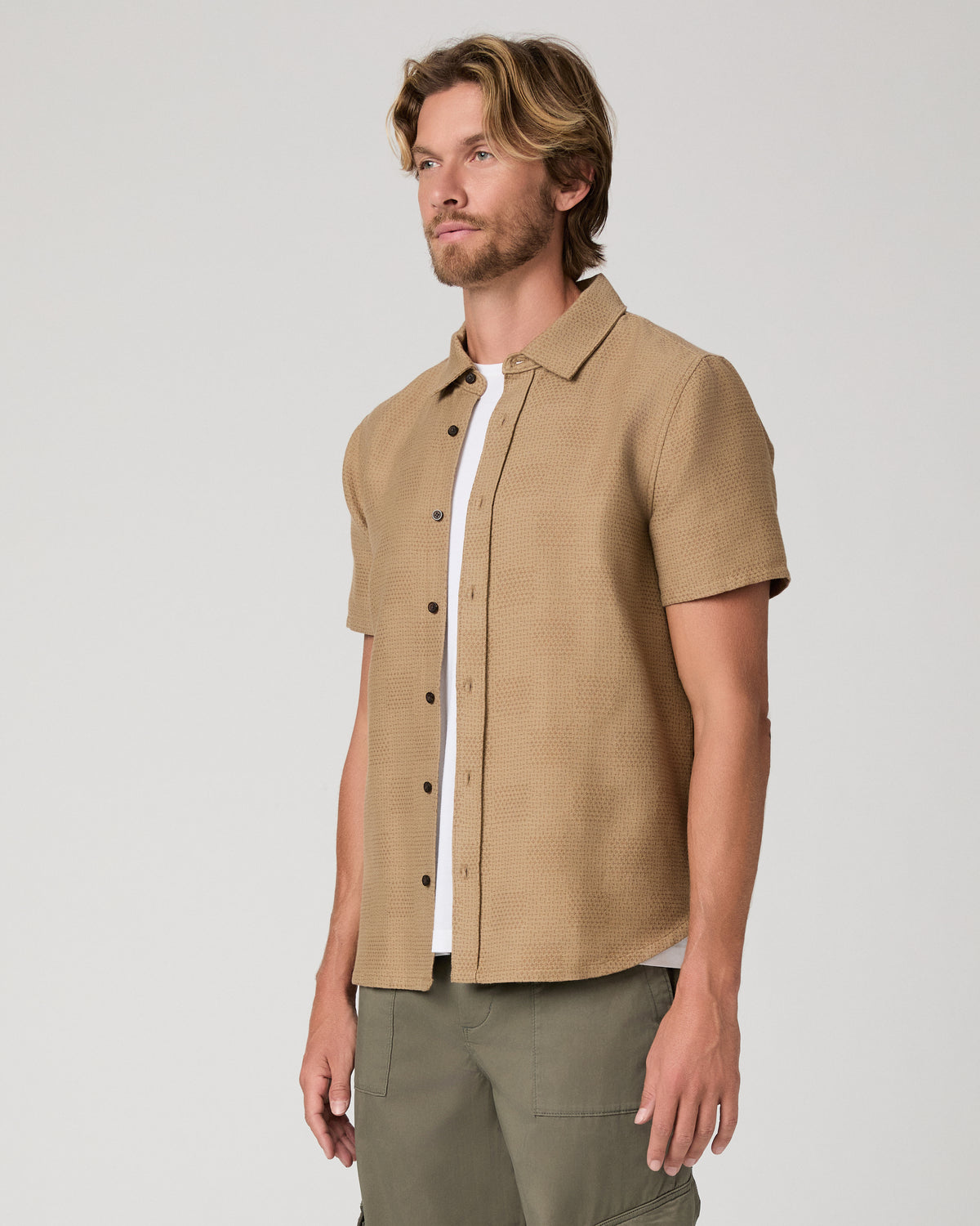 A man wearing a tan short-sleeve button-up shirt over a white t-shirt and olive green pants, standing against a light gray studio background.
