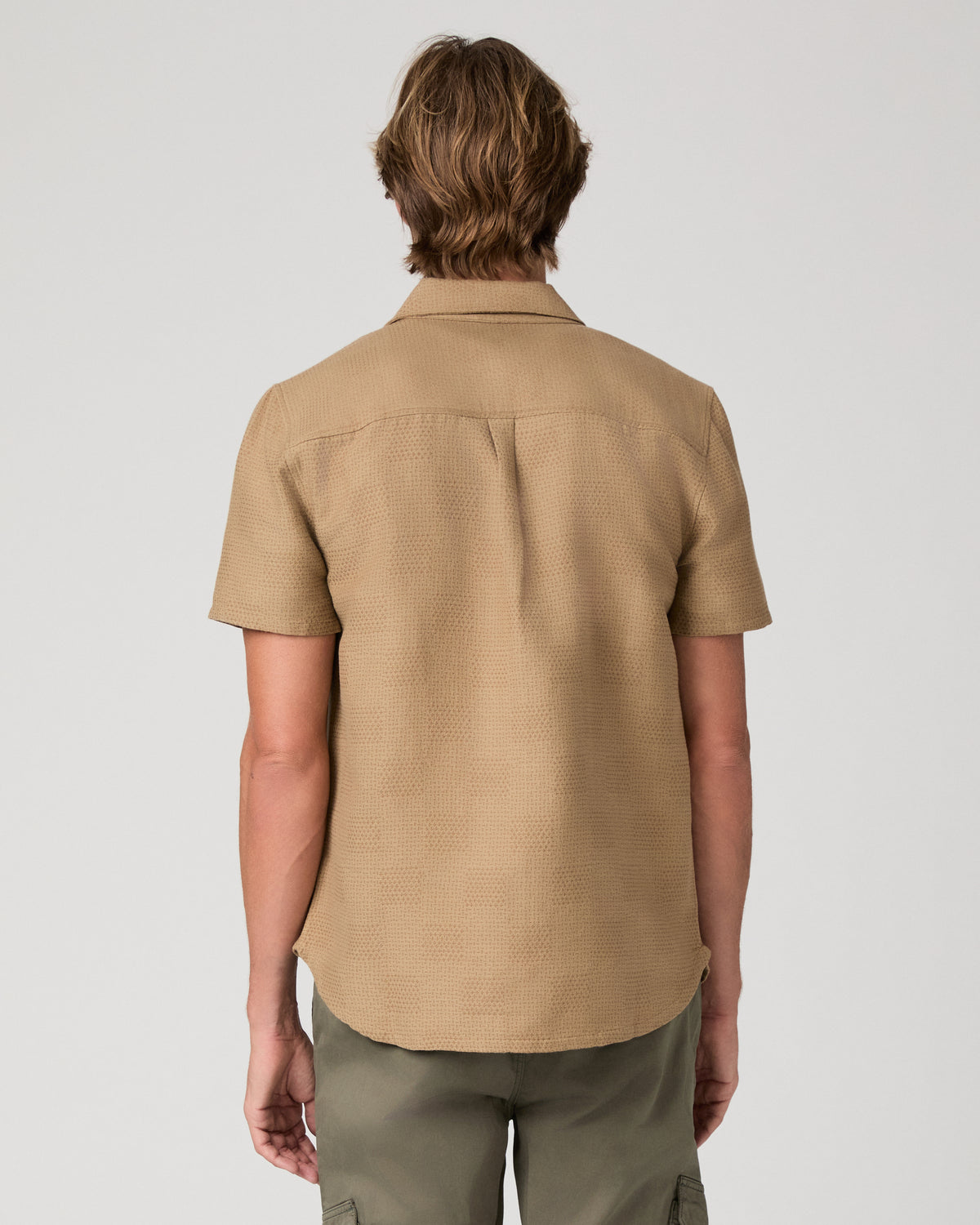 A man wearing a tan short-sleeve button-up shirt over a white t-shirt and olive green pants, standing against a light gray studio background.