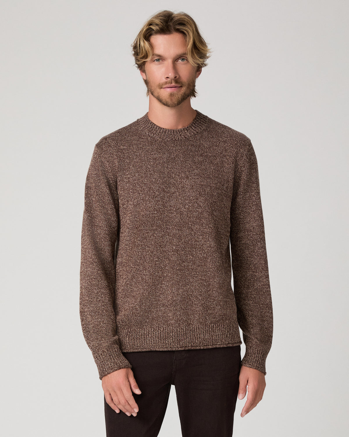 A man wearing a brown knit crewneck sweater and dark pants, standing against a light gray studio background.
