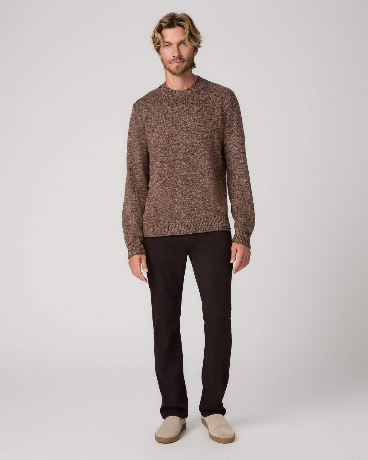 A man wearing a brown knit crewneck sweater and dark pants, standing against a light gray studio background.