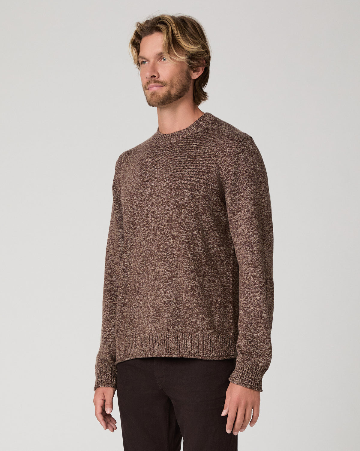 A man wearing a brown knit crewneck sweater and dark pants, standing against a light gray studio background.