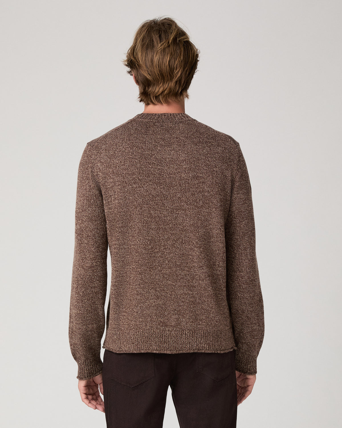 A man wearing a brown knit crewneck sweater and dark pants, standing against a light gray studio background.
