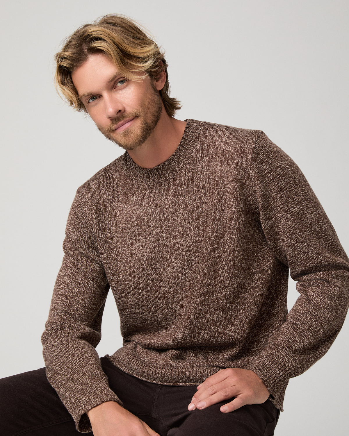 A man wearing a brown knit crewneck sweater and dark pants, standing against a light gray studio background.