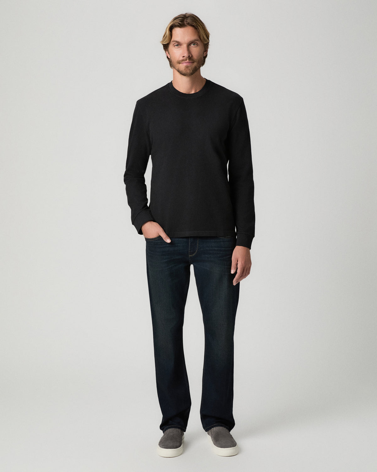 Full-length view of model with one hand in his pocket, wearing the Norcross Long Sleeve Boxy Tee in Vintage Black with dark-wash jeans and grey slip-on shoes
