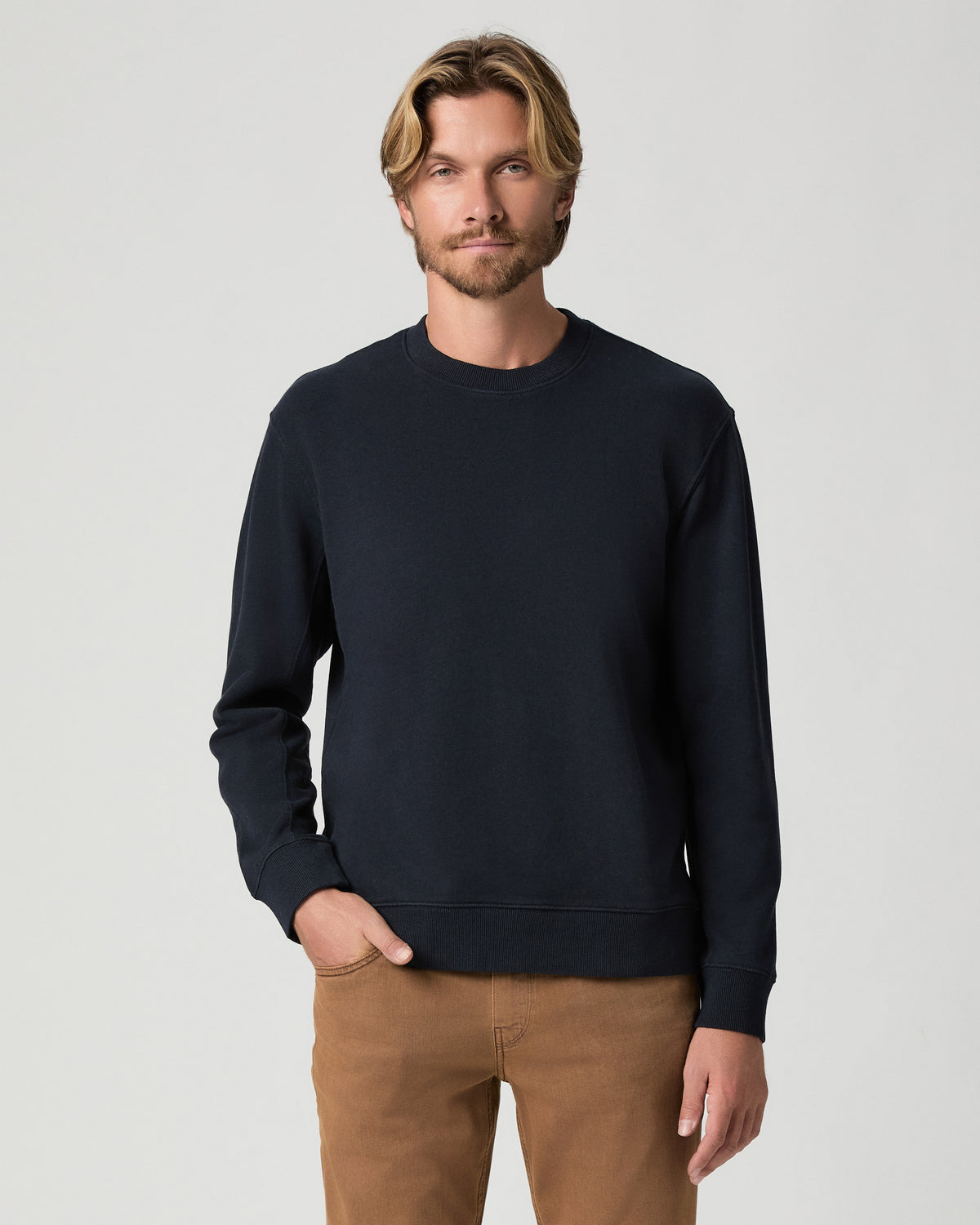 Model standing with one hand in his pocket, wearing the Devlin Crewneck in Deep Anchor, a navy crewneck sweatshirt, with tan pants