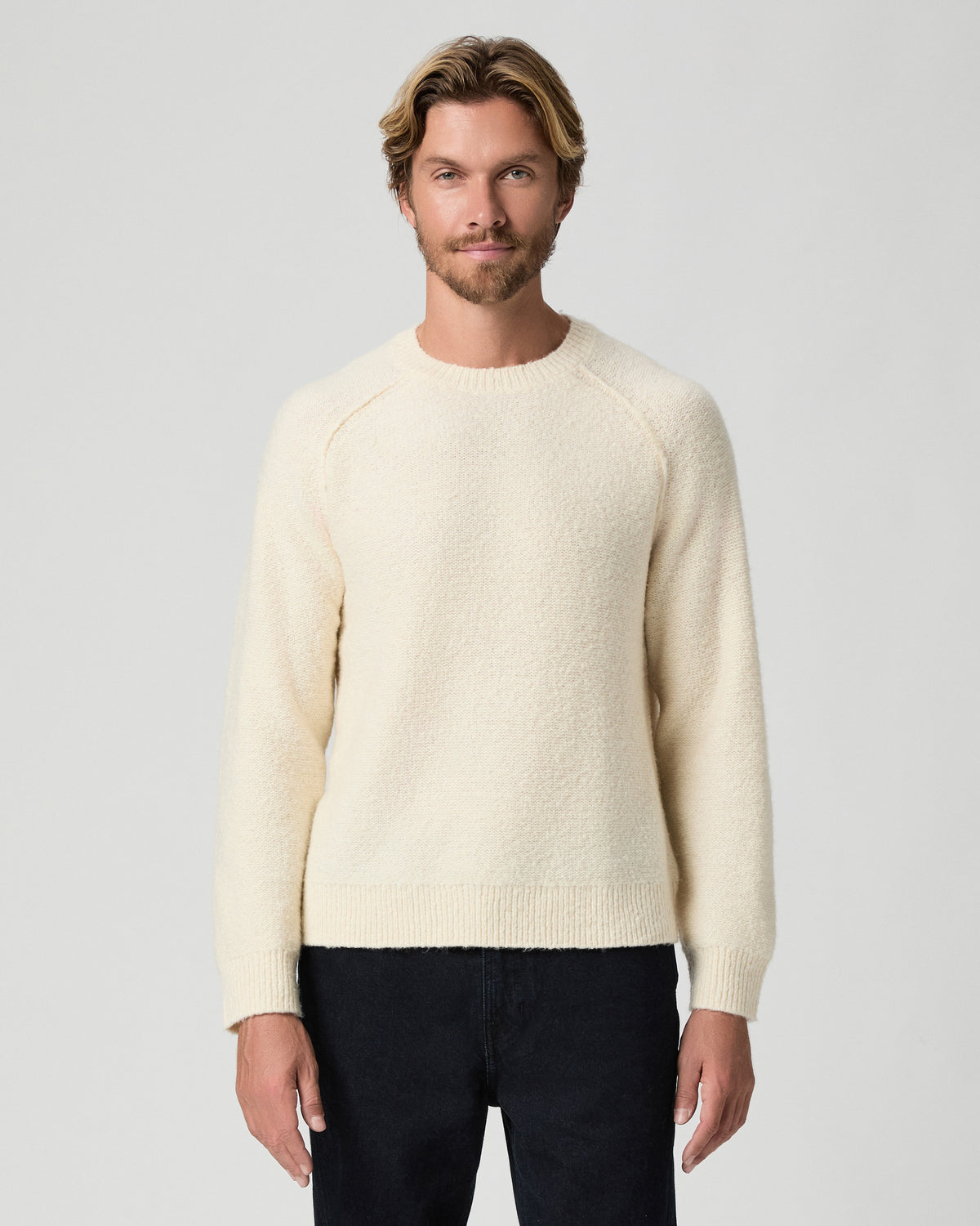 Model standing with arms at his sides, wearing the Hightower Crewneck Sweater in Marfil, a cream knit sweater, with dark jeans