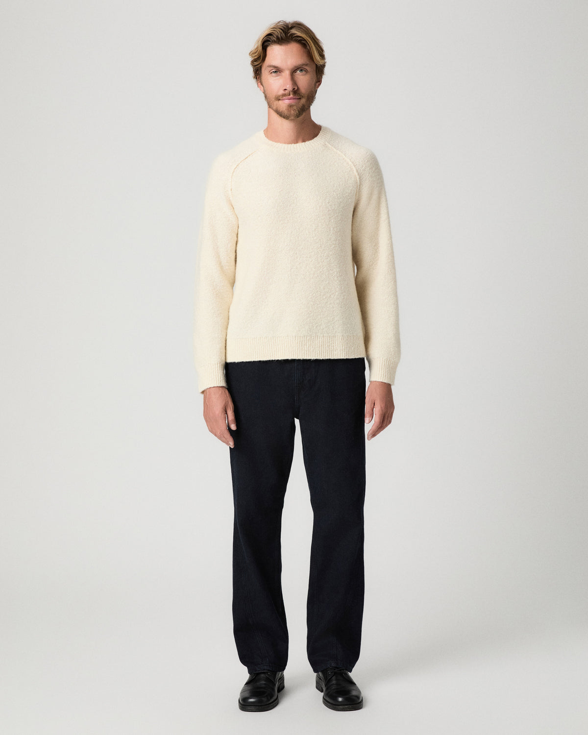 Full-length view of model standing with arms at his sides, wearing the Hightower Crewneck Sweater in Marfil with dark relaxed jeans and black boots